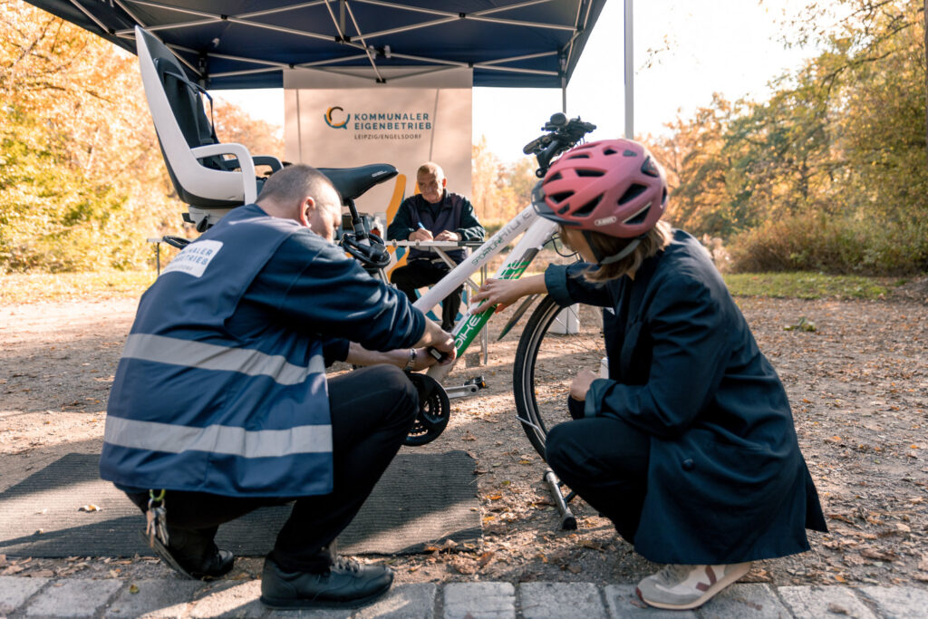 Ein Mann und eine Frau hocken auf dem Boden vor einem Fahrrad. Im Hintergrund sitzt noch ein Mann und es ist der Schriftzug "Kommunaler Eigenbetrieb Leipzig/Engelsdorf" zu sehen.